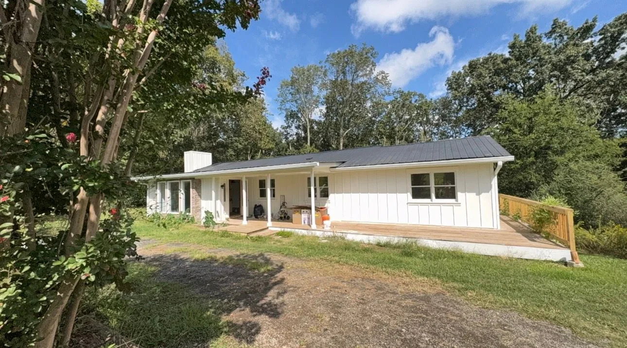 Front exterior of B-504 Young Loop in Fairmount with a white single-story home, covered porch, deck, lawn, gravel drive, and wooded surroundings