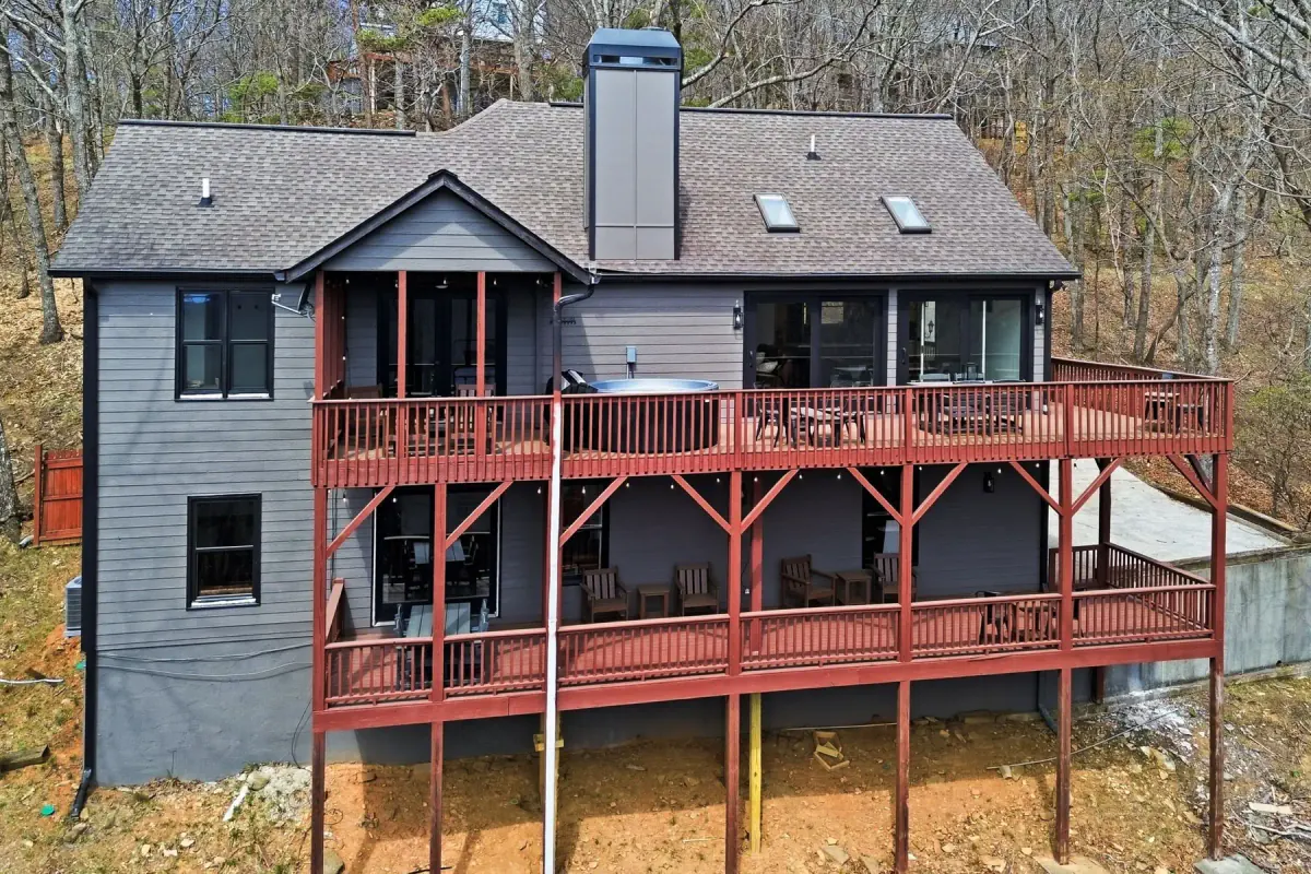 Two-story dark-sided mountain home with two red wood decks, a hot tub, and wooded surroundings